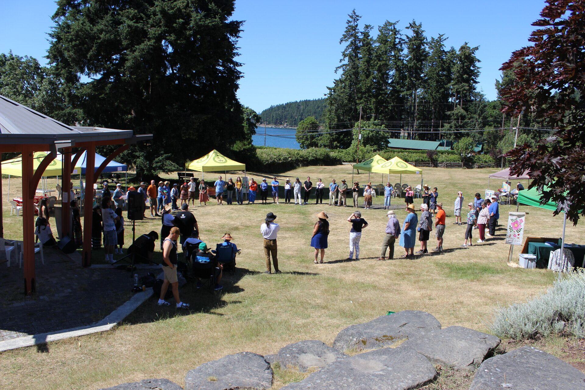 People standing in a wide circle on a grassy field overlooking water and forested shore on SḴŦAḴ / Mayne Island. Temporary Home place photograph.